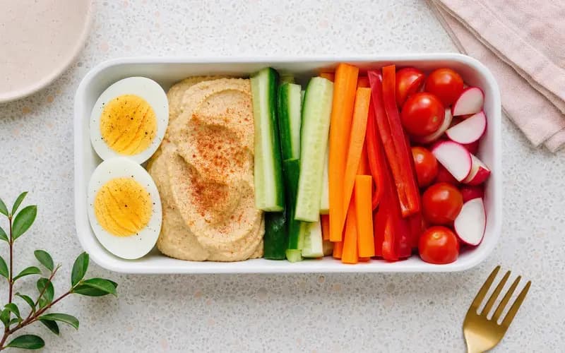 Box repas avec œufs durs coupés en deux, houmous au paprika et bâtonnets de crudités colorées