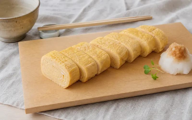 Tamagoyaki tranché en rouleaux épais sur planche en bois clair, texture satinée jaune pâle avec légères couches visibles, radis daikon râpé à côté